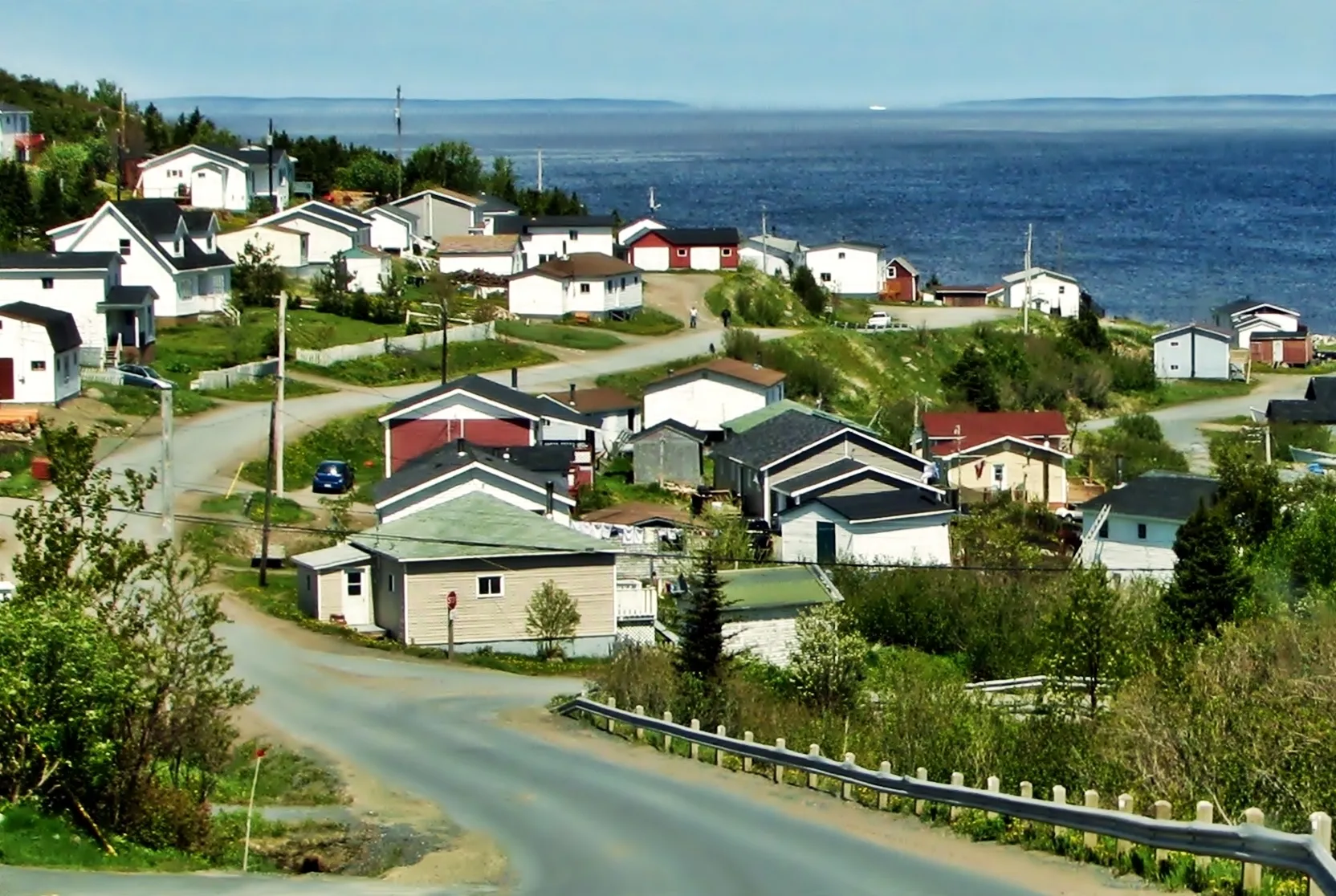Houses stepping down a hillside road to the sea on the Baie Verte Peninsula, open water and distant headlands beyond.