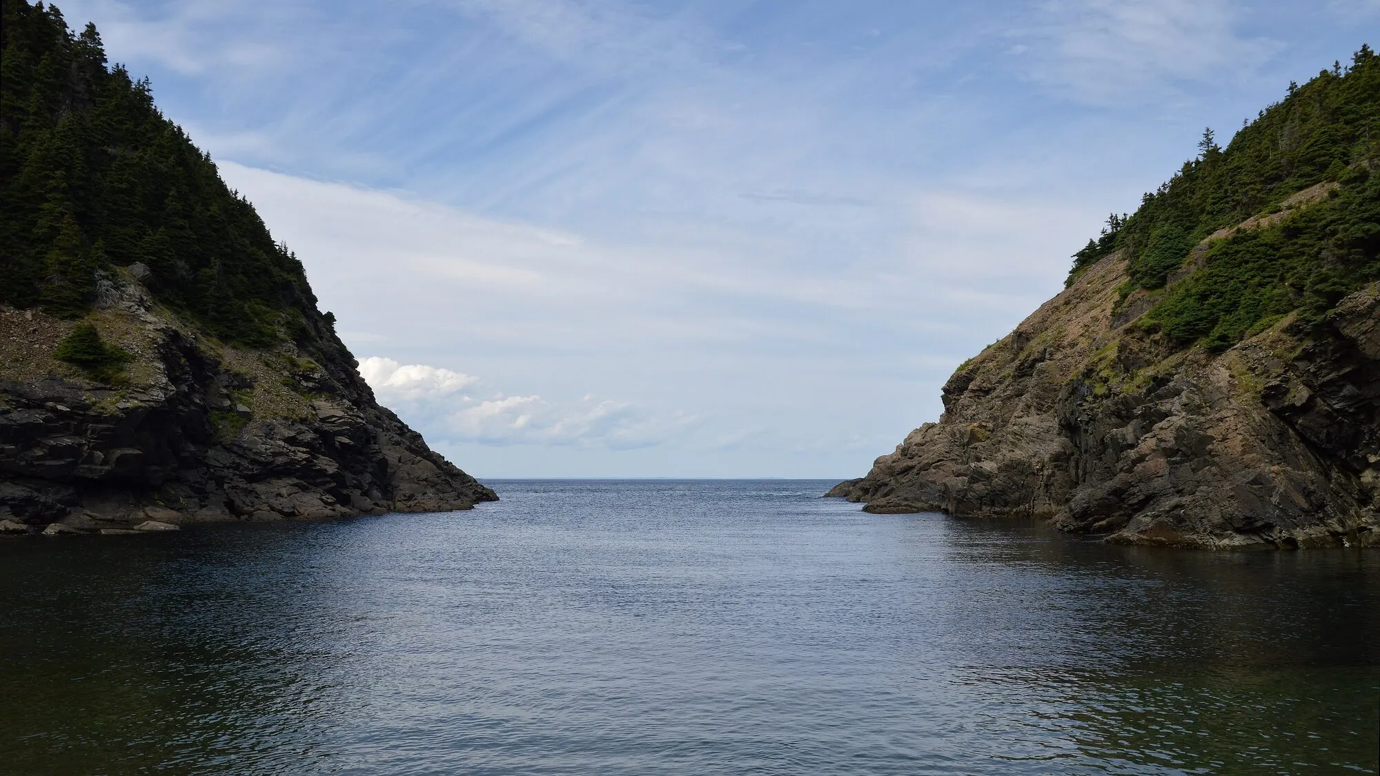 A cove at Pouch Cove between two wooded cliffs, calm water opening to the sea.