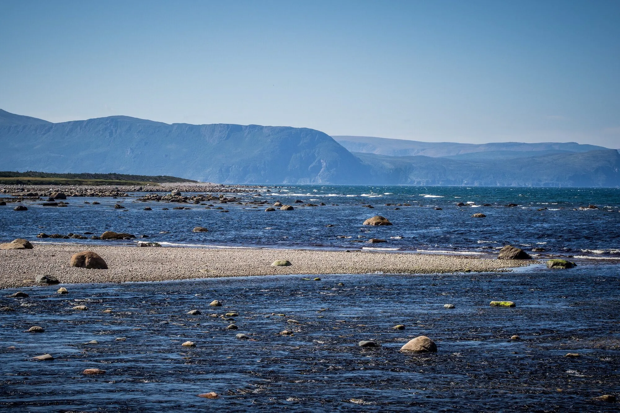Rocks and shingle in the blue sea, with mountains across the water.