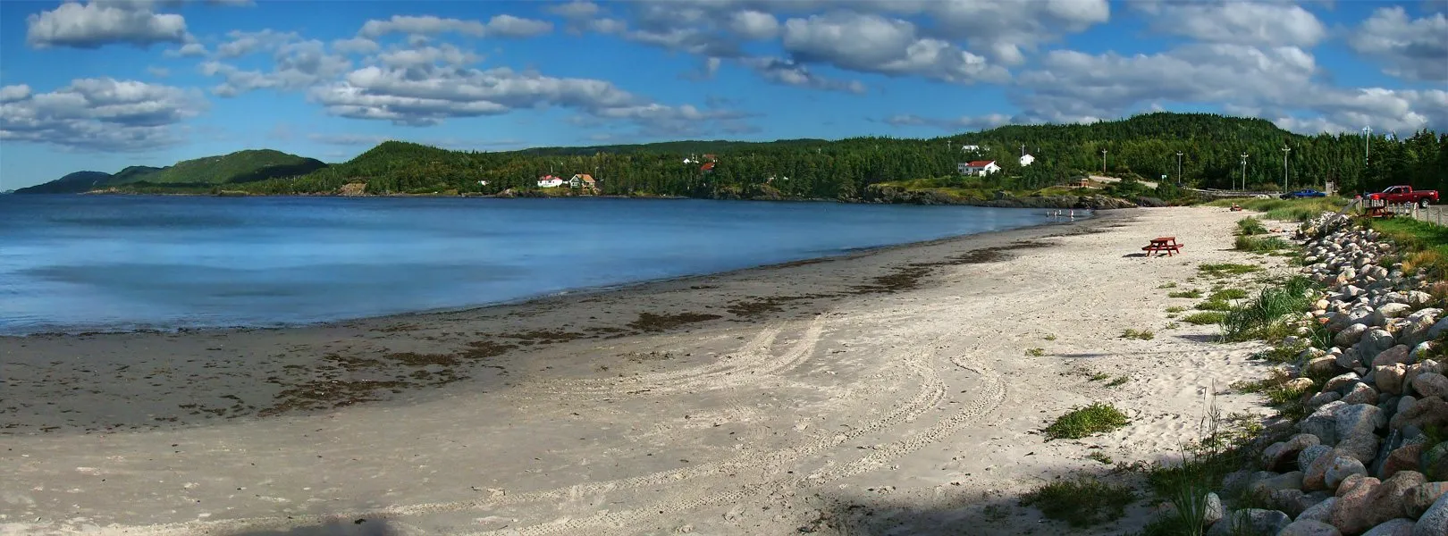 A long sandy beach at Eastport curving along a calm bay, with picnic tables, boulders and wooded hills behind.
