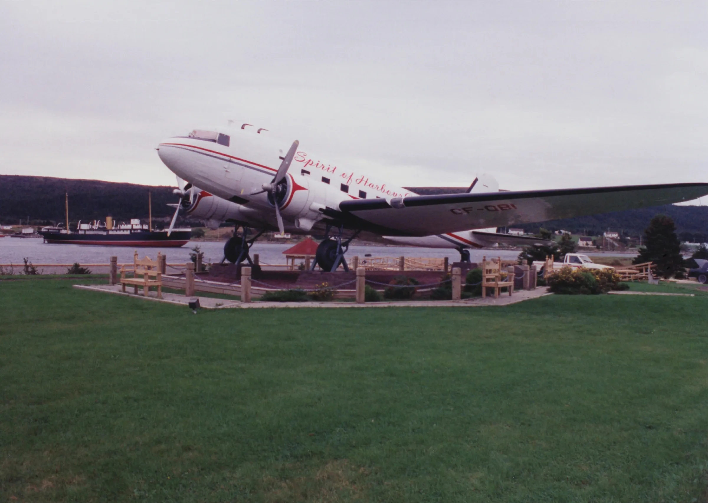 The Spirit of Harbour Grace, a DC-3 mounted beside the water in the town, with the SS Kyle aground across the harbour behind it.