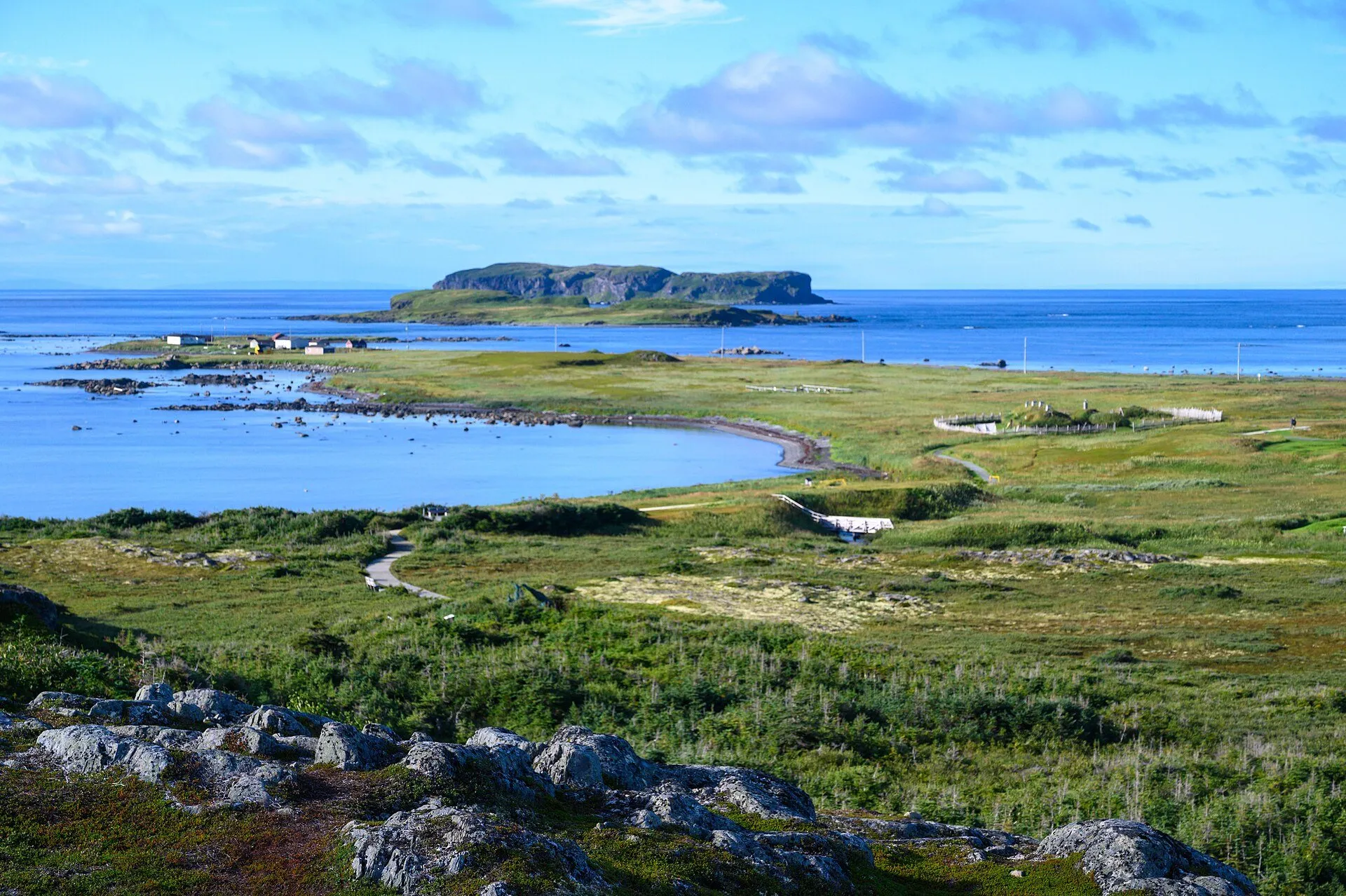 The view from the ridge at L'Anse aux Meadows over green meadows, a curving bay and a flat-topped island offshore.