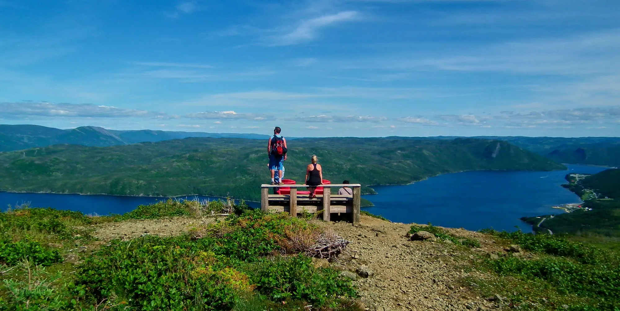 Two hikers at the bench on top of the Lookout Trail, looking out over Bonne Bay and forested hills.