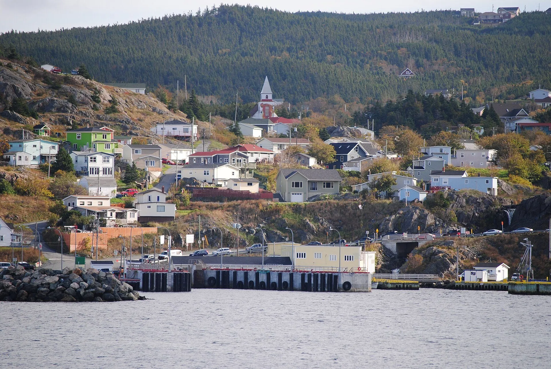 The Portugal Cove ferry wharf at the water's edge, with houses and a church climbing the wooded hill behind.