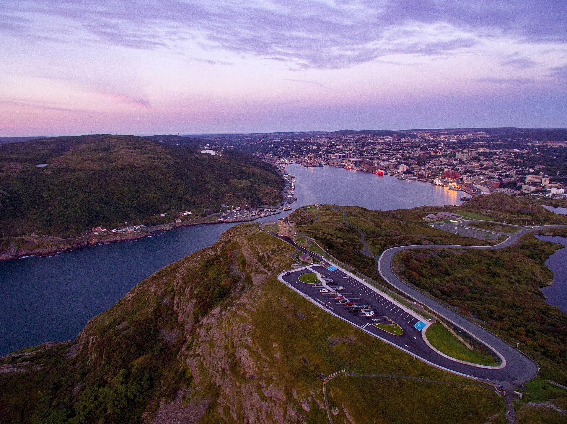 Signal Hill from the air at dusk: Cabot Tower on the summit above the Narrows, the road winding up past the car park, and St. John's harbour and city behind.