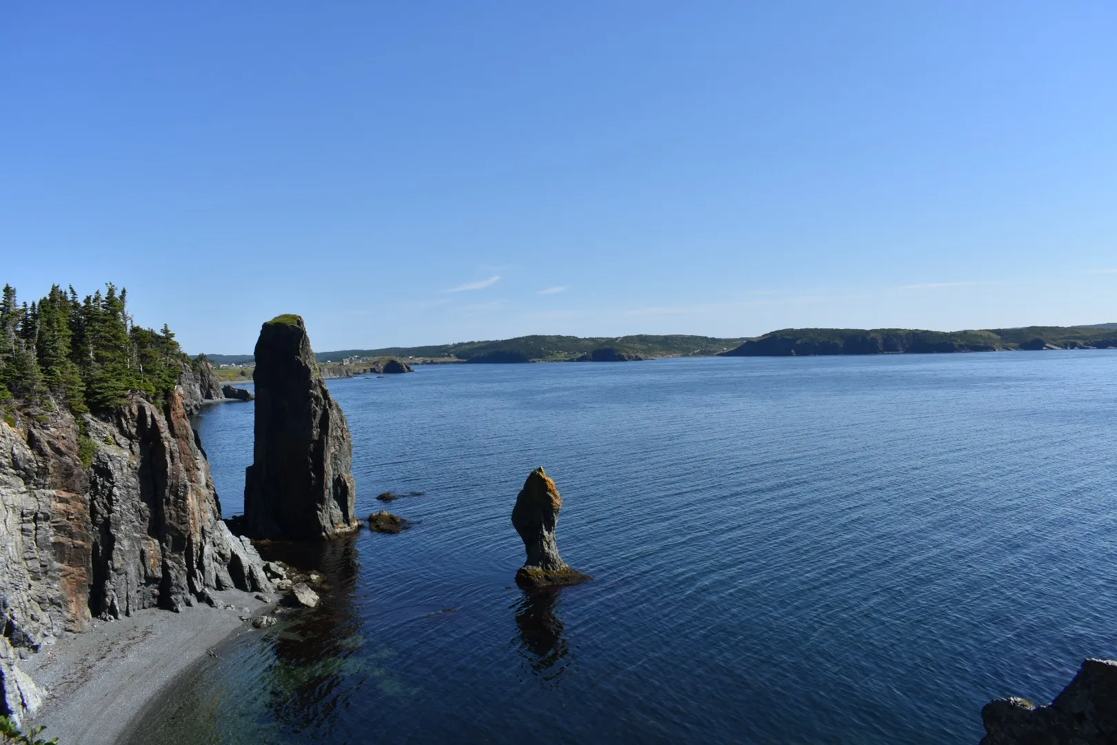 Two tall sea stacks standing off a cliff and a grey stone beach on the Skerwink Trail at Port Rexton, the far shore of Trinity Bay behind.