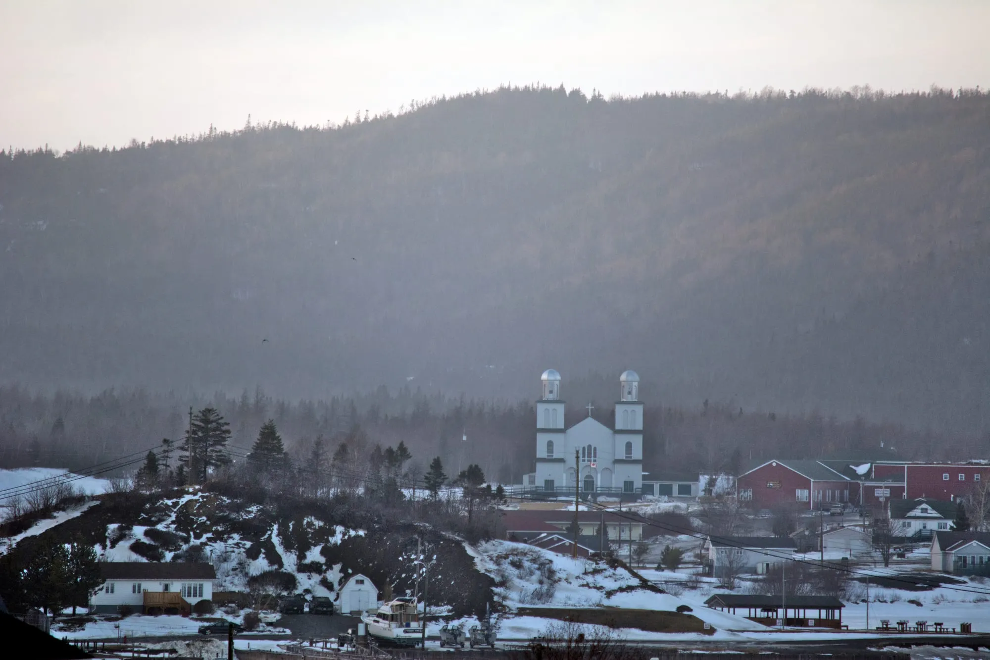 St. Alban's in winter, the white church of St. Ignatius Parish above the houses, a dark wooded hill behind.