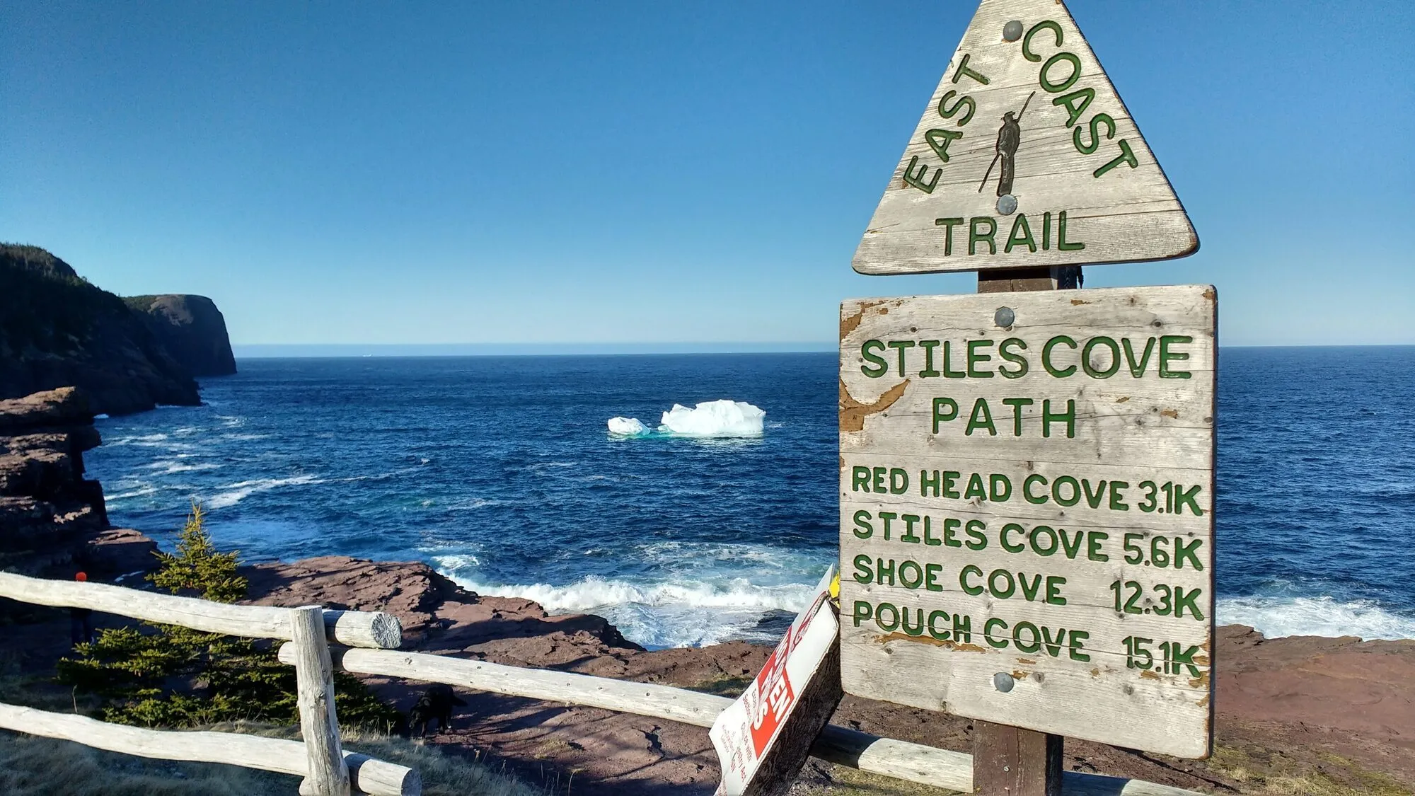 The East Coast Trail's Stiles Cove Path sign beside a rail fence above the sea, an iceberg offshore.