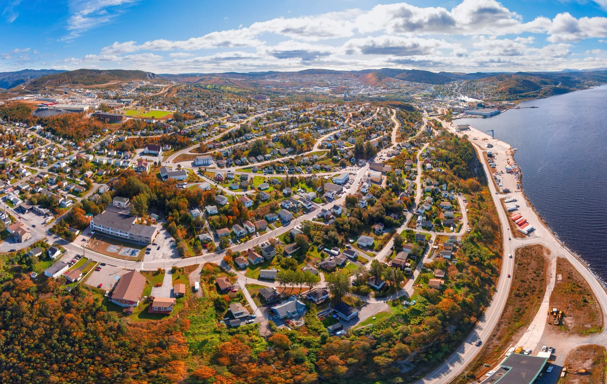Aerial of Corner Brook in autumn colour along the Humber Arm shore, hills behind, blue sky with cloud.