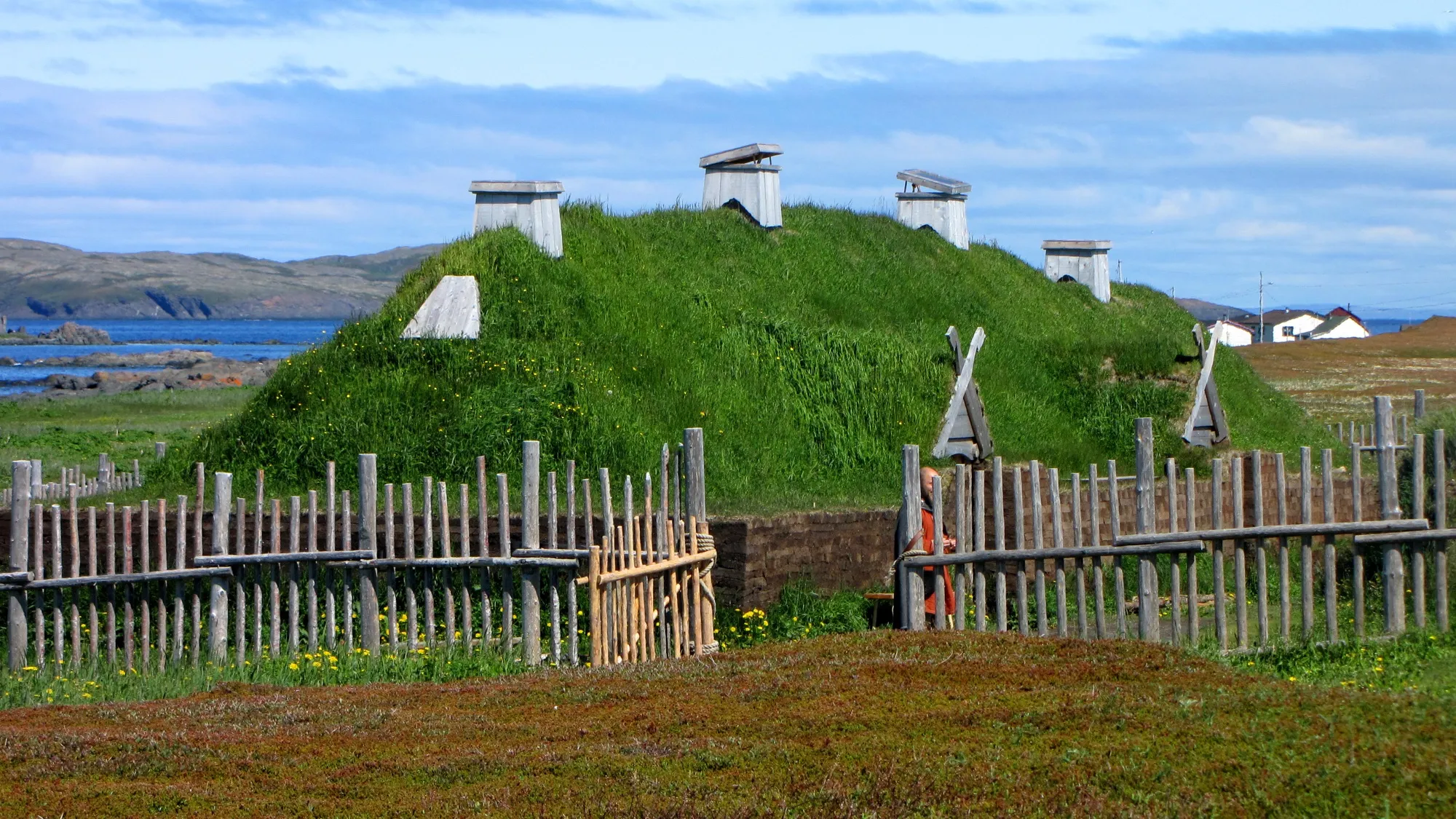 The recreated Norse sod longhouse behind a palisade, sea and coast behind, blue sky.