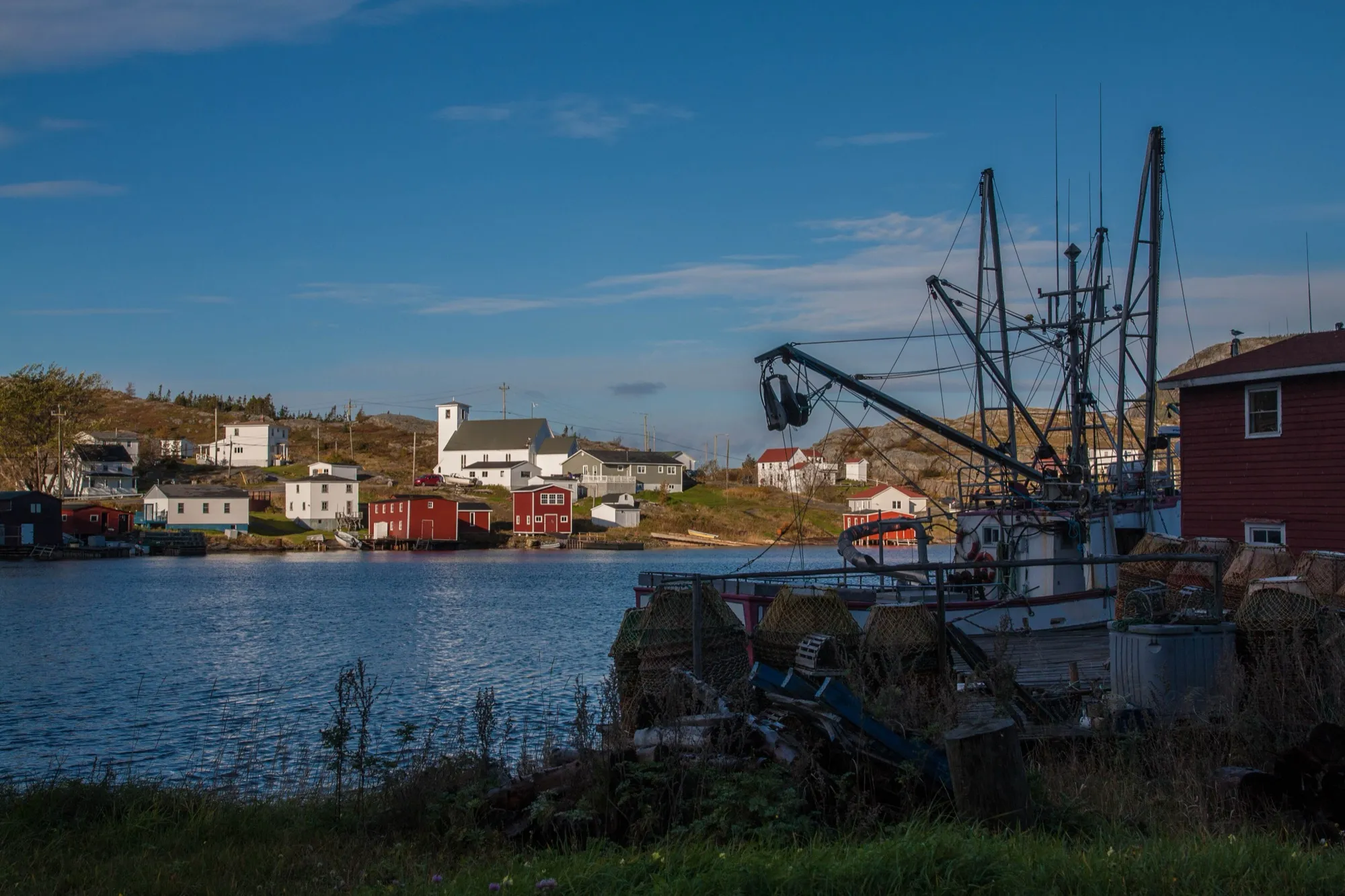 Salvage harbour in evening sun: church, red stages and houses across the water, longliner and lobster pots in foreground.